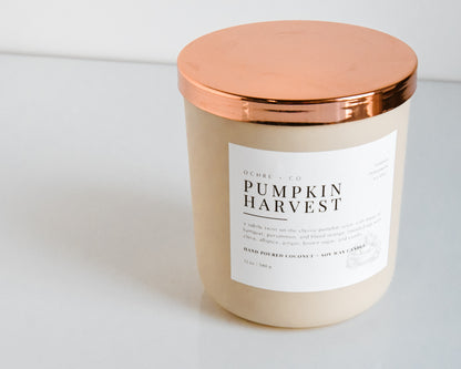 A matte beige candle vessel with a copper lid and a label that reads "Pumpkin Harvest" on a white background