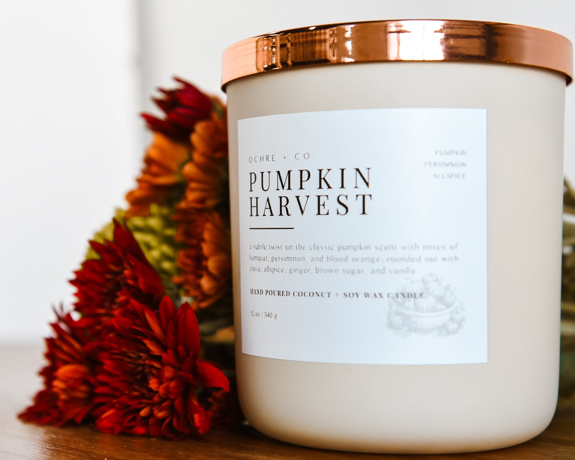 A matte beige ceramic candle vessel with a copper lid and a label that reads "Pumpkin Harvest" on a wooden counter and fall-colored florals
