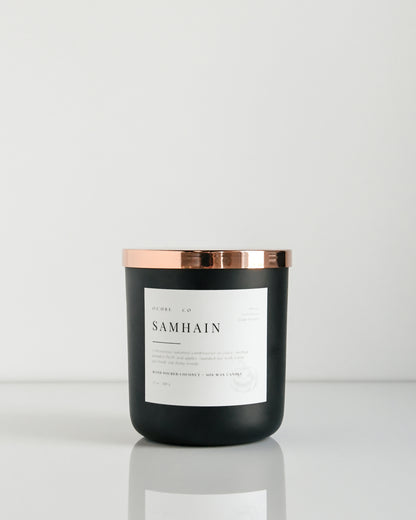 A matte black candle with a copper lid labeled 'Samhain' set against a white background.