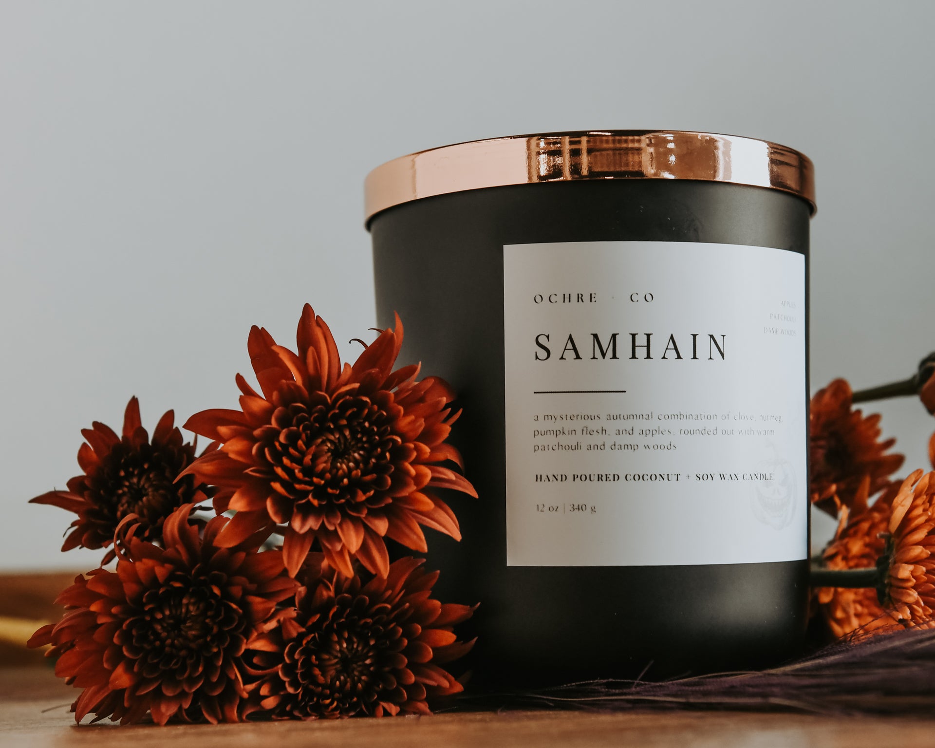 A matte black candle with a copper lid labeled 'Samhain' set against a white background on a wood countertop and beautiful orange florals