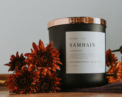 A matte black candle with a copper lid labeled 'Samhain' set against a white background on a wood countertop and beautiful orange florals