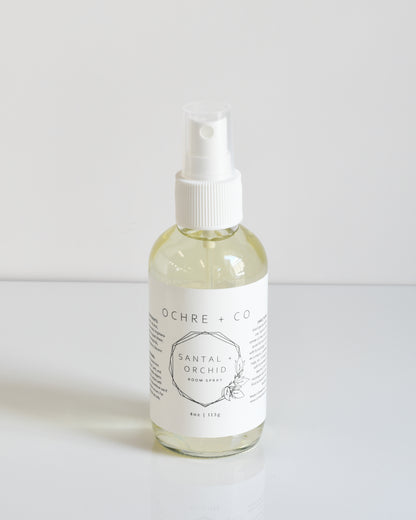 A clear glass bottle with a white spray nozzle labeled 'Santal + Orchid Room Spray' on a white background