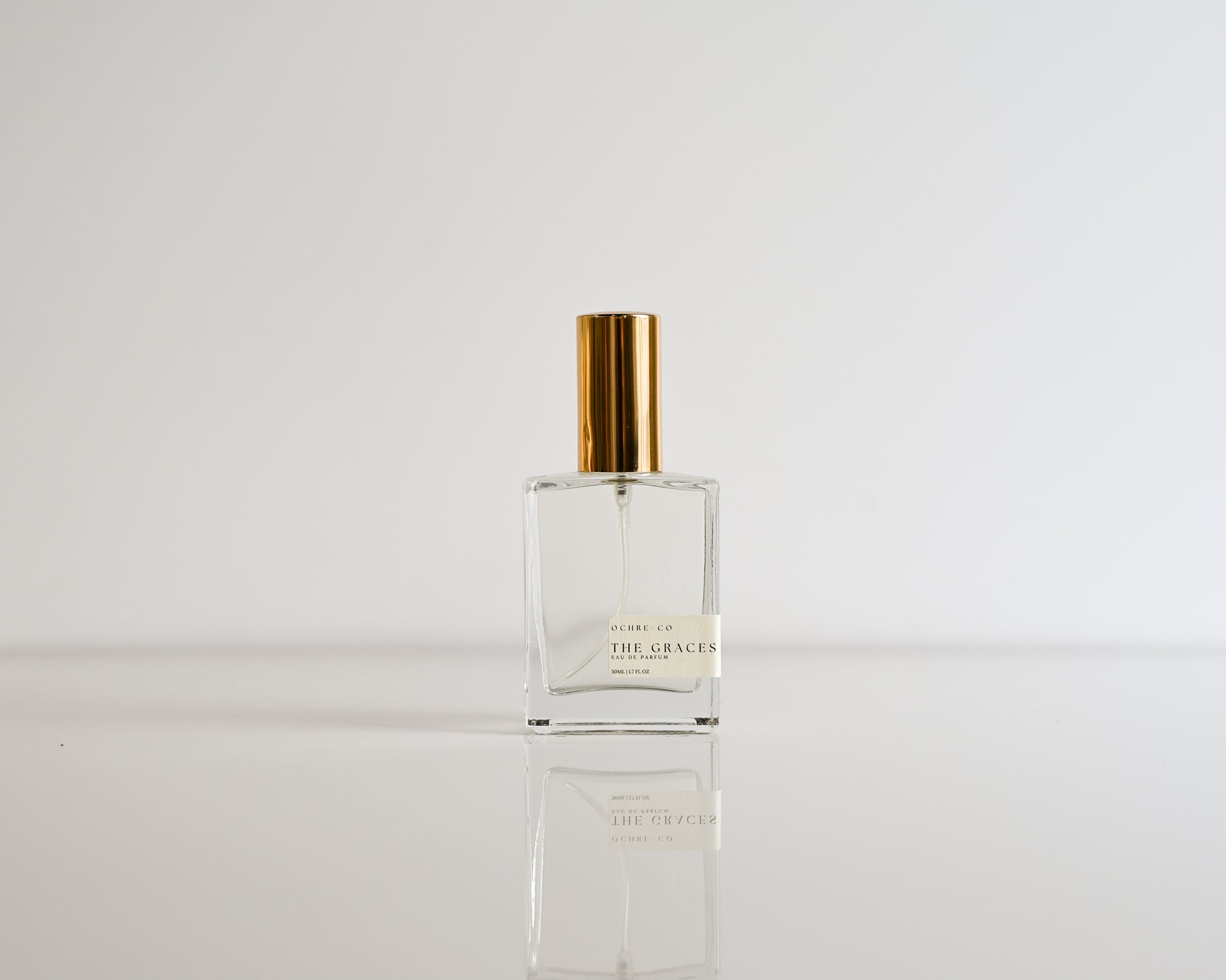 Clear perfume bottle of The Graces with gold cap on a white background