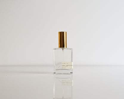 Clear perfume bottle of The Graces with gold cap on a white background