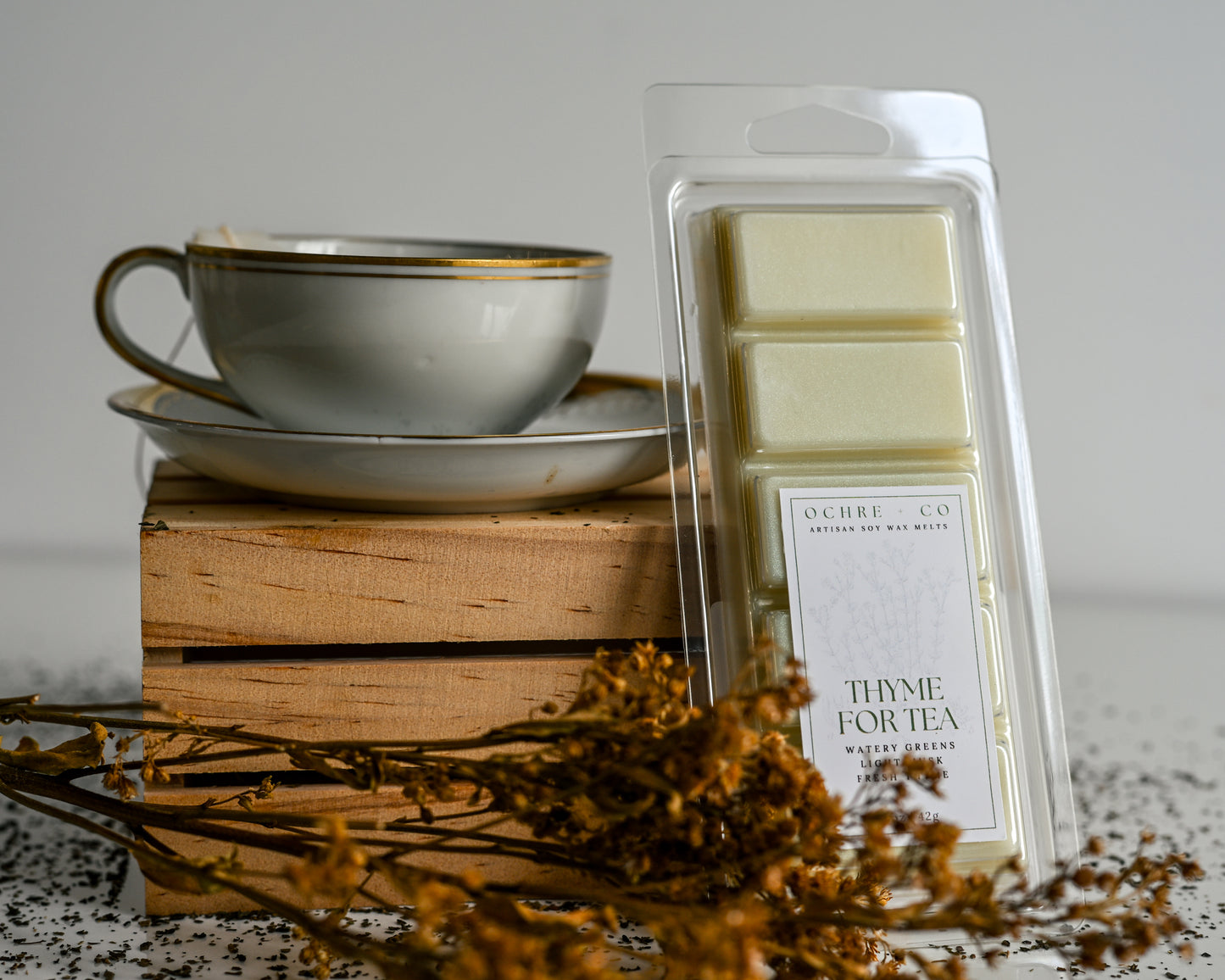 Thyme for Tea wax melt on a white background and reflective surface with scattered tea leaves soft orange florals, and a tea cup