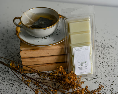 Thyme for Tea wax melt on a white background and reflective surface with scattered tea leaves soft orange florals, and a tea cup
