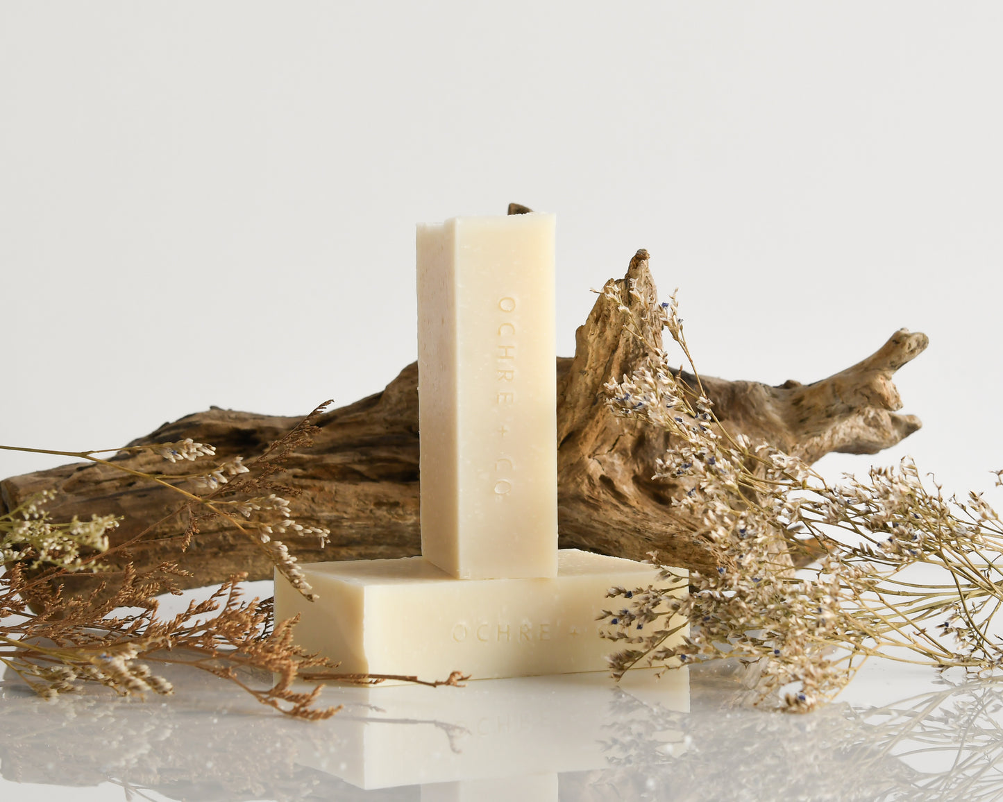 Two bars of soap on a white surface with dried plants and a piece of driftwood.