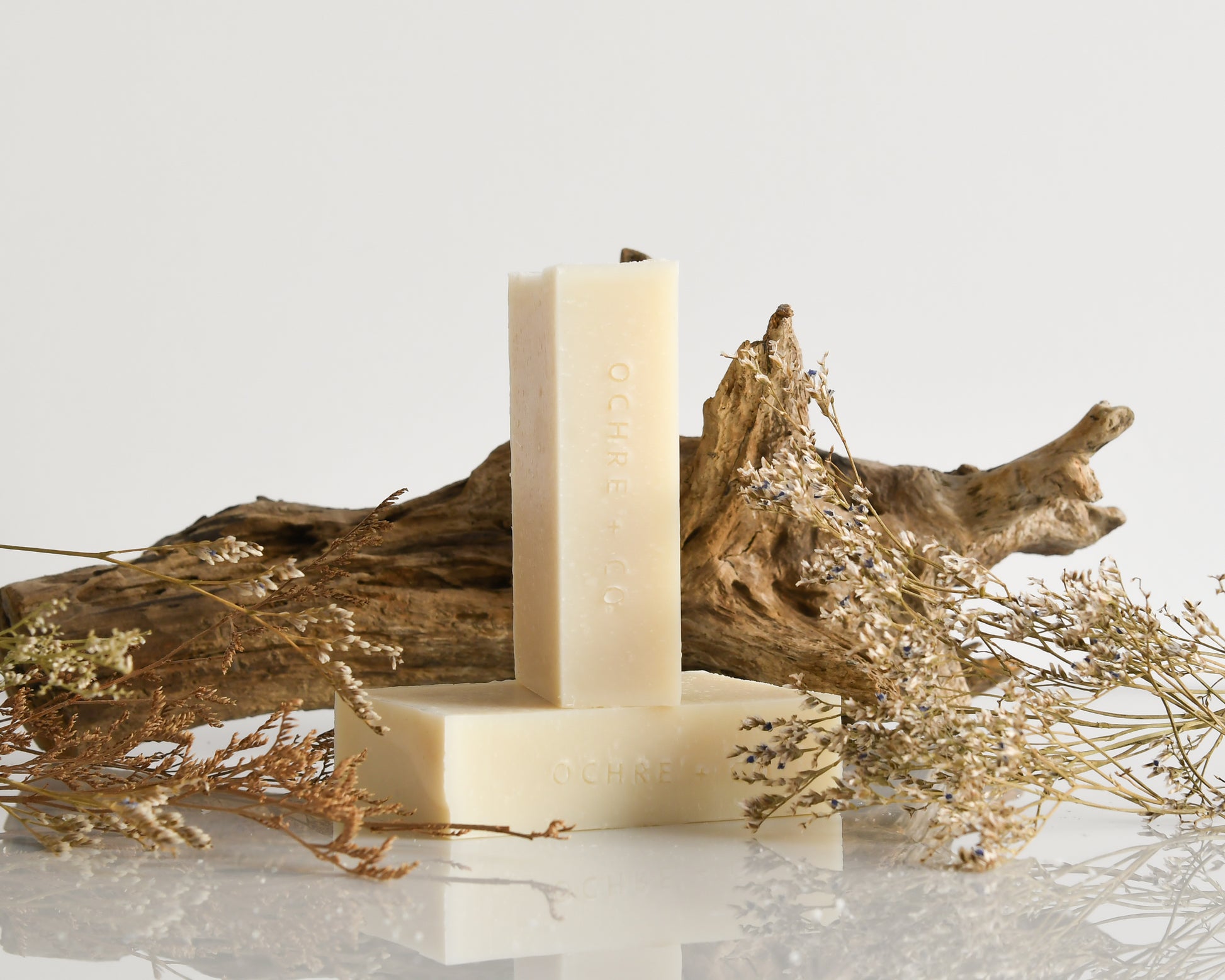 Two bars of soap on a white surface with dried plants and a piece of driftwood.