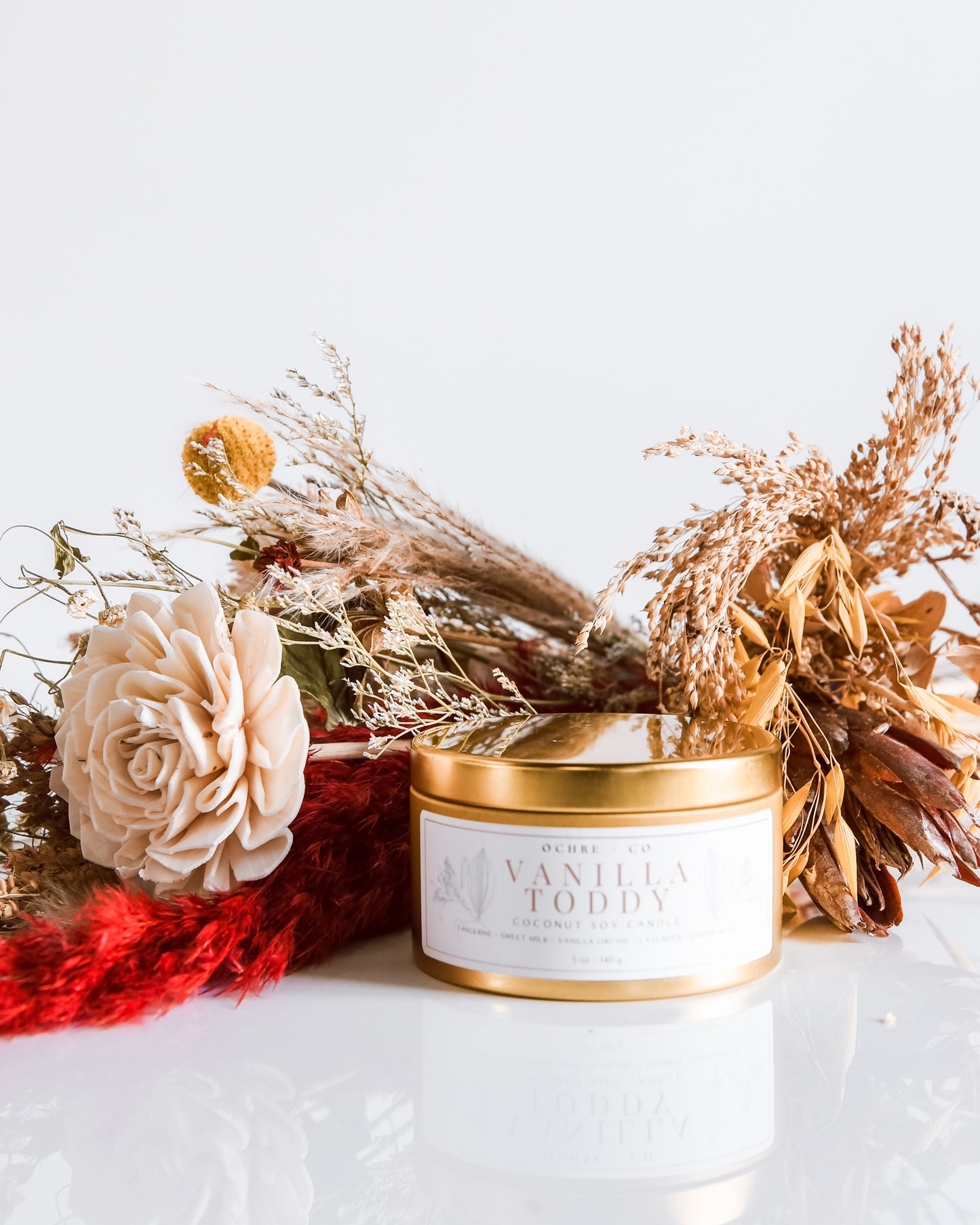 A gold metal candle vessel with a label, surrounded by decorative items including a red textured object and dried flowers.